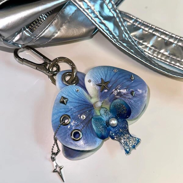 以下が含まれることがあります： 青い蘭の花の形をしたキーホルダー。銀色の星型スタッズ、小さなラインストーン、中央の白いパール、チェーンで吊るされた銀色の星のチャームが付いています。銀色のバッグにナスカンで取り付けられています。