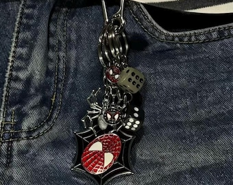 Op de afbeelding: Een metalen sleutelhanger met een rood-zwarte spinnenwebbedel met een maskerembleem, een zilveren spinnenbedel, een kleine rood-zwarte maskerbedel en twee zeszijdige dobbelstenen in zwart en grijs, bevestigd aan een zilverkleurige metalen ring.