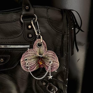 Op de afbeelding: Een zwarte leren handtas met een decoratieve orchideeënbedel. De bedel is een paarse en witte orchidee met zilveren piercings en een ketting met een sterbedel. De tas heeft een rits, metalen hardware en franjes.