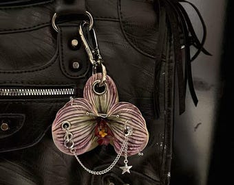 Op de afbeelding: Een zwarte leren handtas met een decoratieve orchideeënbedel. De bedel is een paarse en witte orchidee met zilveren piercings en een ketting met een sterbedel. De tas heeft een rits, metalen hardware en franjes.