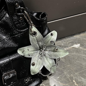 Op de afbeelding: Een zwarte handtas met textuur en een grote, lichtgroene, lelievormige bedel die is bevestigd met een zilverkleurige metalen clip. De bloemblaadjes zijn versierd met kleine zilveren studs en ringetjes, en er hangt een fijne zilveren ketting aan.