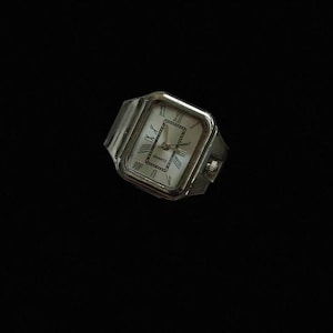 Op de afbeelding: Een zilverkleurig rechthoekig quartz polshorloge met een witte wijzerplaat, zwarte Romeinse cijfers en een decoratieve rand in het midden. Het horloge heeft een metalen kast, een kroon aan de zijkant en geïntegreerde bandaanzetten.