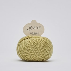 May include: A ball of light olive green cashmere yarn with a beige paper label attached to the top. The label features the text Cardiff Cashmere and the word classic printed in black, along with a small graphic of a goat silhouette.