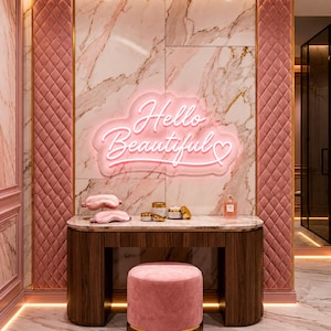 Op de afbeelding: Een roze make-up tafel set. Het bevat een houten make-up tafel met een marmeren blad, een roze gestoffeerde kruk en een neonbord met de tekst "Hello Beautiful". De muren zijn versierd met roze gewatteerde panelen en marmer.