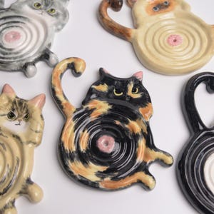 Op de afbeelding: Een verzameling van vijf keramische zeepbakjes in de vorm van een kat, elk met een uniek vachtpatroon zoals lapjeskat, cyperse kat en effen zwart. De bakjes hebben een cirkelvormig midden met concentrische ribbels en een klein roze detail.