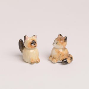 Op de afbeelding: Twee miniatuur keramieken kattenbeeldjes die naast elkaar zitten. De linker kat heeft een crèmekleurige vacht met een donkerbruin gezicht, oren en staart. De rechter kat heeft oranje, witte en zwarte cyperpatronen met een pluizige staart.