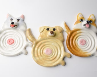 May include: Three ceramic dog-shaped trinket dishes featuring a white dog, a golden dog, and a brown and white dog. Each dish has a circular, rippled center with a small pink accent and a sculpted dog head and tail extending from the rim.