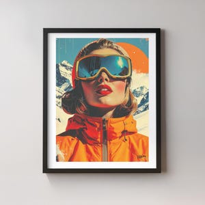 May include: Framed art print featuring a woman wearing ski goggles and an orange jacket, set against a backdrop of snow-capped mountains and a vibrant orange sun. The artwork has a vintage, retro style.