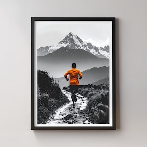 Pode incluir: Impressão emoldurada em preto e branco de um corredor em uma trilha de montanha. O corredor está vestindo uma camisa laranja, criando um contraste marcante com a paisagem em tons de cinza. Uma montanha coberta de neve domina o fundo, adicionando profundidade à cena.