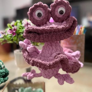 May include: A handmade, whimsical crochet creature in shades of pink and mauve. The plush toy features large, googly eyes and a ruffled, textured design. The creature has a unique, playful appearance, perfect for a child's room or as a decorative item.