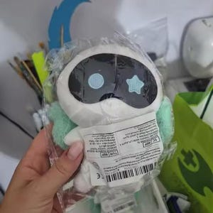 May include: A white plush toy in a clear plastic bag. The toy has a black oval head with two blue circles and a star shape. It has light green accents and is held by a hand.