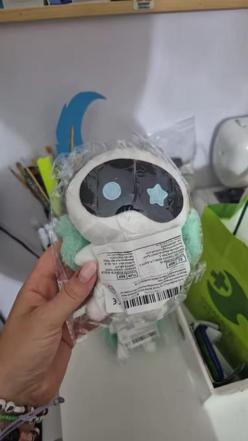 May include: A white plush toy in a clear plastic bag. The toy has a black oval head with two blue circles and a star shape. It has light green accents and is held by a hand.