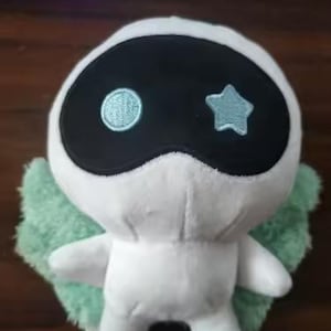 May include: A white plush toy with a black mask-like face, featuring a light blue circle and star. It has light green wings and small arms. The toy is designed to resemble a character.