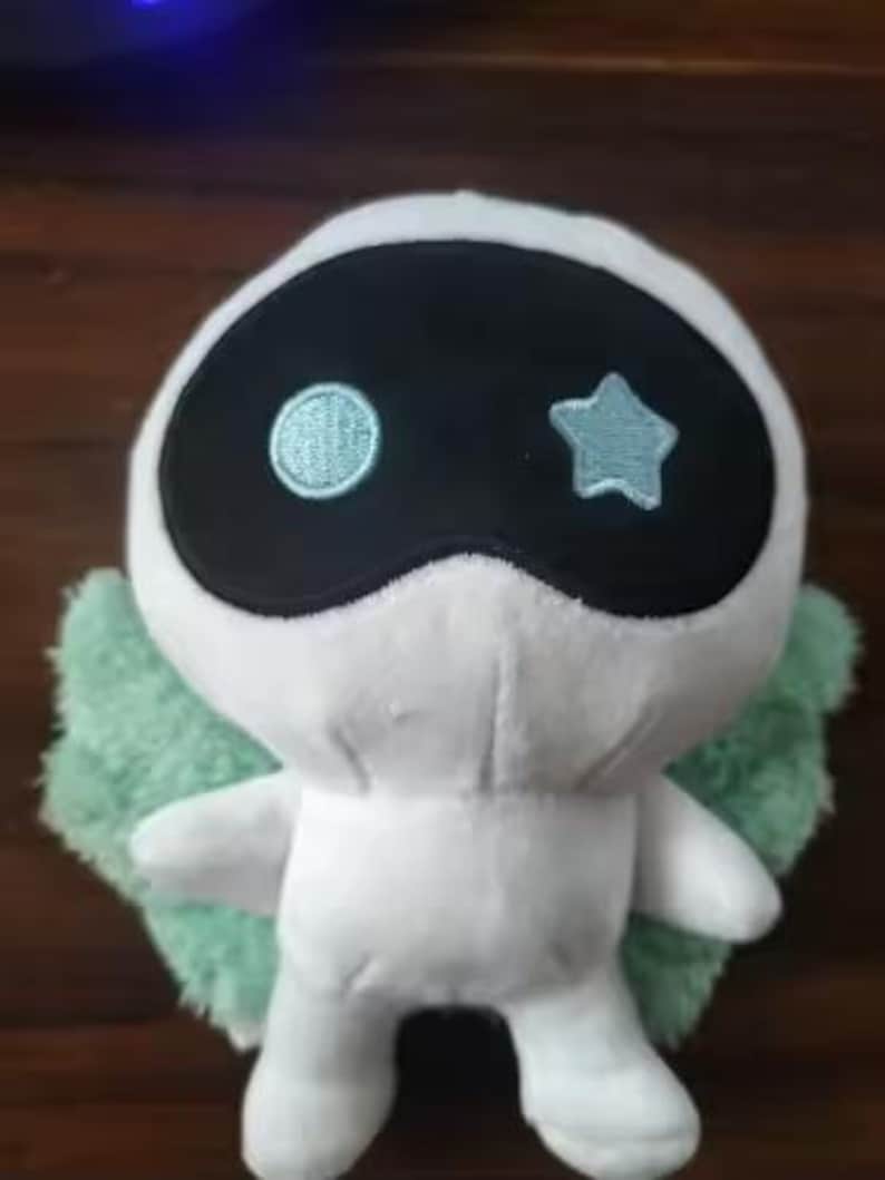 May include: A white plush toy with a black mask-like face, featuring a light blue circle and star. It has light green wings and small arms. The toy is designed to resemble a character.