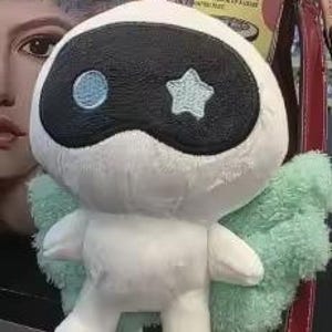 May include: A white plush toy with a black mask-like design and a blue star and circle. It has light green wings and is attached to a keychain. The toy is hanging near a floral patterned bag and a red strap.