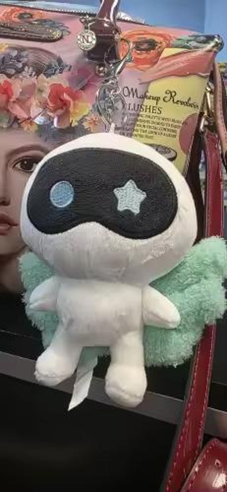 May include: A white plush toy with a black mask-like design and a blue star and circle. It has light green wings and is attached to a keychain. The toy is hanging near a floral patterned bag and a red strap.