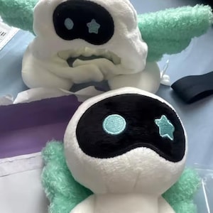May include: Two plush toys, one a hat and the other a doll, both featuring a white base with black eye-shaped masks. The hat has mint green wings, and the doll has mint green wings and small arms. Both have a blue circle and star detail.