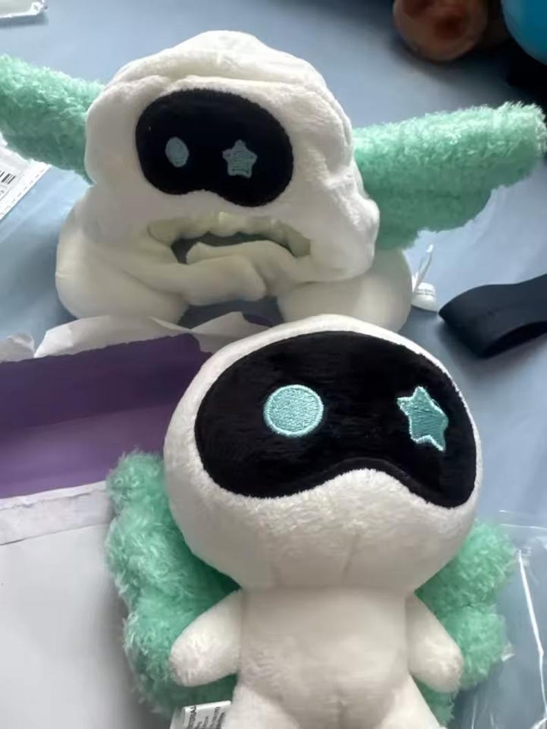 May include: Two plush toys, one a hat and the other a doll, both featuring a white base with black eye-shaped masks. The hat has mint green wings, and the doll has mint green wings and small arms. Both have a blue circle and star detail.