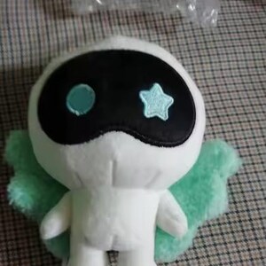 May include: A white plush toy with a black mask-like face, featuring a blue circle and a star. It has light green wings and is set against a checkered background. The toy is a cute, whimsical character.