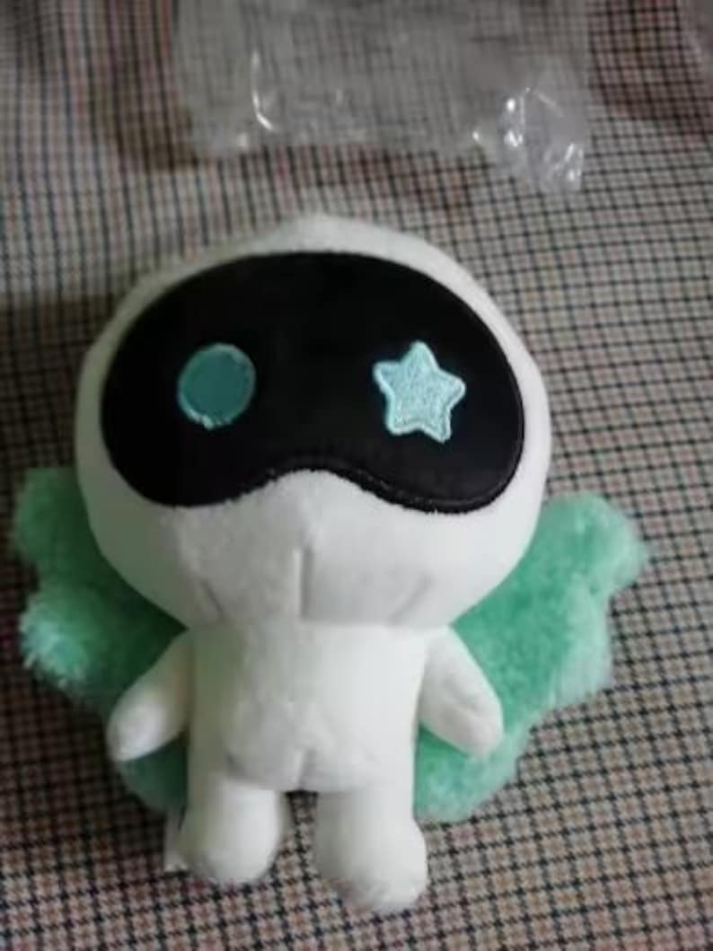 May include: A white plush toy with a black mask-like face, featuring a blue circle and a star. It has light green wings and is set against a checkered background. The toy is a cute, whimsical character.