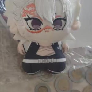 May include: A plush doll of a character with white hair and a black and white outfit. The doll has a detailed face with purple eyes and a scar-like design on the forehead. The doll is sitting on a surface with coins.