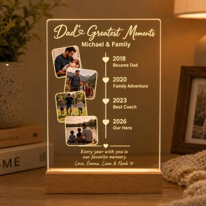 May include: A personalized LED light with a wooden base. The acrylic panel displays family photos and text: "Dad's Greatest Moments" with the names "Michael & Family". It includes dates and captions like "Became Dad" and "Our Hero".