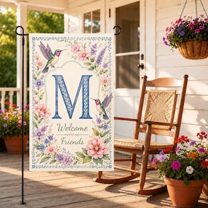 May include: A garden flag with a floral design and the letter "M". The flag features pink, purple, and blue flowers, two hummingbirds, and the words "Welcome Friends". The flag is displayed on a black metal stand.