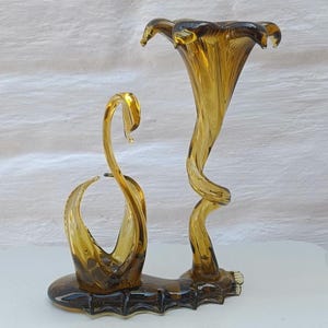 May include: Amber-colored glass sculpture featuring a stylized flower and a swan-like form. The flower has a flared, ruffled top and a spiraling stem. The swan-like form has a curved neck and a small, open bowl. The sculpture sits on a dark brown base.