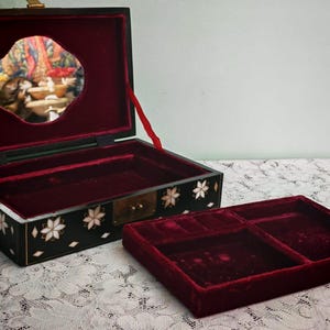 May include: A black rectangular jewelry box with a hinged lid, featuring white floral mother-of-pearl inlays on the exterior. The interior is lined with deep red velvet and includes a removable tray with compartments and a mirror in the lid.