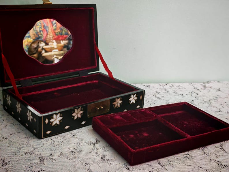May include: A black rectangular jewelry box with a hinged lid, featuring white floral mother-of-pearl inlays on the exterior. The interior is lined with deep red velvet and includes a removable tray with compartments and a mirror in the lid.