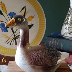 May include: A ceramic duck figurine with a brown and white body, a black head, and a yellow beak. The duck is resting on a wooden stand. A decorative plate with a yellow rim and floral design is in the background.