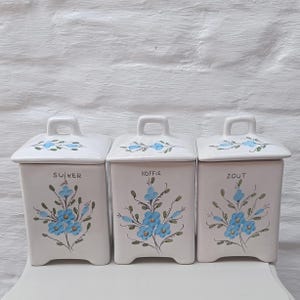 May include: Three white ceramic canisters with lids, each labeled with a different word. The canisters are decorated with blue floral designs and have small handles on the lids. The words on the canisters are "SUJKER", "KOFFIE", and "ZOUT".