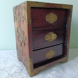 Könnte beinhalten: Eine kleine Holzkommode mit drei Schubladen. Die Kommode hat eine dunkelrotbraune Farbe mit Messing-Ecken und Schubladengriffen. Die Seiten haben dekorative Messingverzierungen.