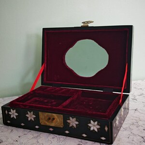 May include: A rectangular black jewelry box with a hinged lid featuring a brass latch. The exterior is decorated with inlaid mother-of-pearl flower and diamond shapes. The interior is lined with deep red velvet and includes a mirror.