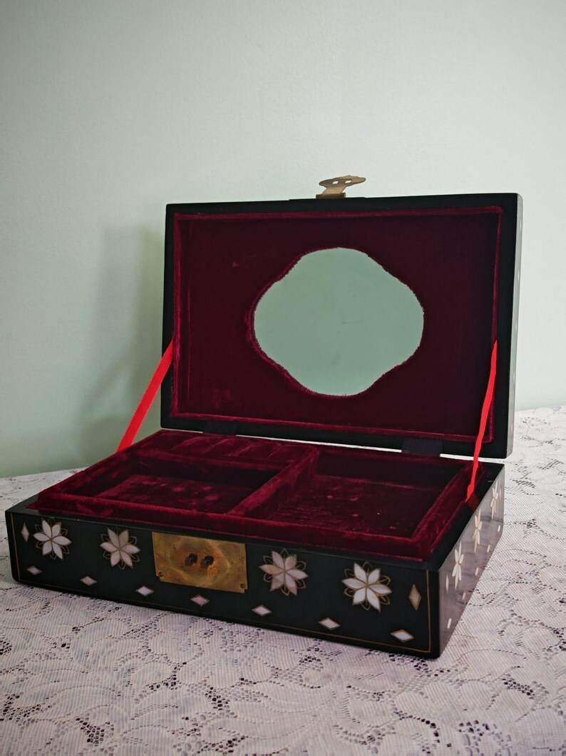May include: A rectangular black jewelry box with a hinged lid featuring a brass latch. The exterior is decorated with inlaid mother-of-pearl flower and diamond shapes. The interior is lined with deep red velvet and includes a mirror.