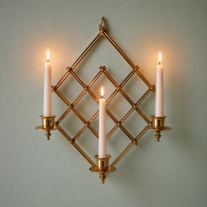Peut inclure: Un bougeoir mural en métal doré doté d'une structure en treillis en forme de losange. L'objet comprend trois coupelles circulaires, chacune supportant une longue bougie effilée blanc cassé avec une flamme allumée, formant un ensemble symétrique.