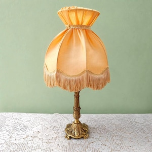May include: A table lamp featuring a gathered peach-coloured fabric shade with a decorative trim and a long fringe along the bottom edge. The lamp has a slender, ornate gold-toned metal base with a textured, fluted design and a wide, circular foot.