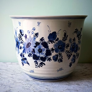 May include: A large, white ceramic planter with a blue floral design. The planter has a wide, rounded shape with a slightly flared rim. The blue floral pattern includes various flower shapes and leaf details. Two blue stripes encircle the top and bottom.