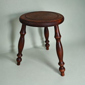 May include: A dark brown wooden stool featuring a circular seat with a visible grain pattern and three turned legs. Each leg has a distinct bulbous shape with rounded sections, providing a stable base for the round top surface of the furniture.
