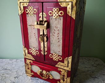 May include: A deep red wooden cabinet featuring two doors with carved white stone panels, brass corner reinforcements, and decorative brass knot motifs. A brass padlock secures the doors above a single drawer with a brass handle and stone inlays.