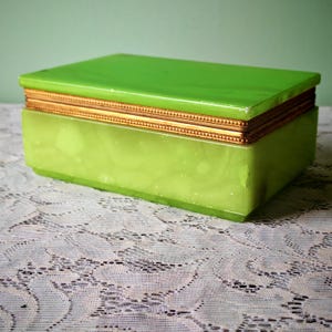 Può includere: Una scatola rettangolare verde lime con finiture dorate. La scatola ha una finitura lucida e poggia su una superficie di pizzo bianco. La scatola viene probabilmente utilizzata per riporre gioielli o piccoli oggetti.