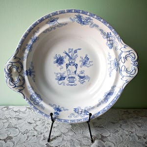 May include: A white ceramic serving dish with blue floral and ribbon patterns. The dish has two handles and a central design of a vase with flowers. The rim and handles are decorated with intricate blue designs. The dish is displayed on a black metal stand.