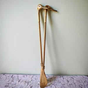 May include: A pair of long-handled brass shoehorns featuring handles shaped like duck heads. The metallic gold-toned implements have slender, straight shafts and wide, curved scoops at the base, designed for assisting with putting on footwear.