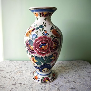 Könnte beinhalten: Eine weiße Keramikvase mit handgemaltem Blumenmuster in Rot-, Blau-, Orange- und Grüntönen. Das Design zeigt große zentrale Blüten, kleinere Akzentblumen und einen Vogel inmitten des Blattwerks, ergänzt durch blaue Bänder am oberen Rand.
