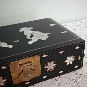 May include: A rectangular black lacquered wooden box featuring inlaid mother-of-pearl figures on the lid and floral motifs on the front. The box includes a prominent brass latch mechanism and thin gold-colored line detailing along the edges.