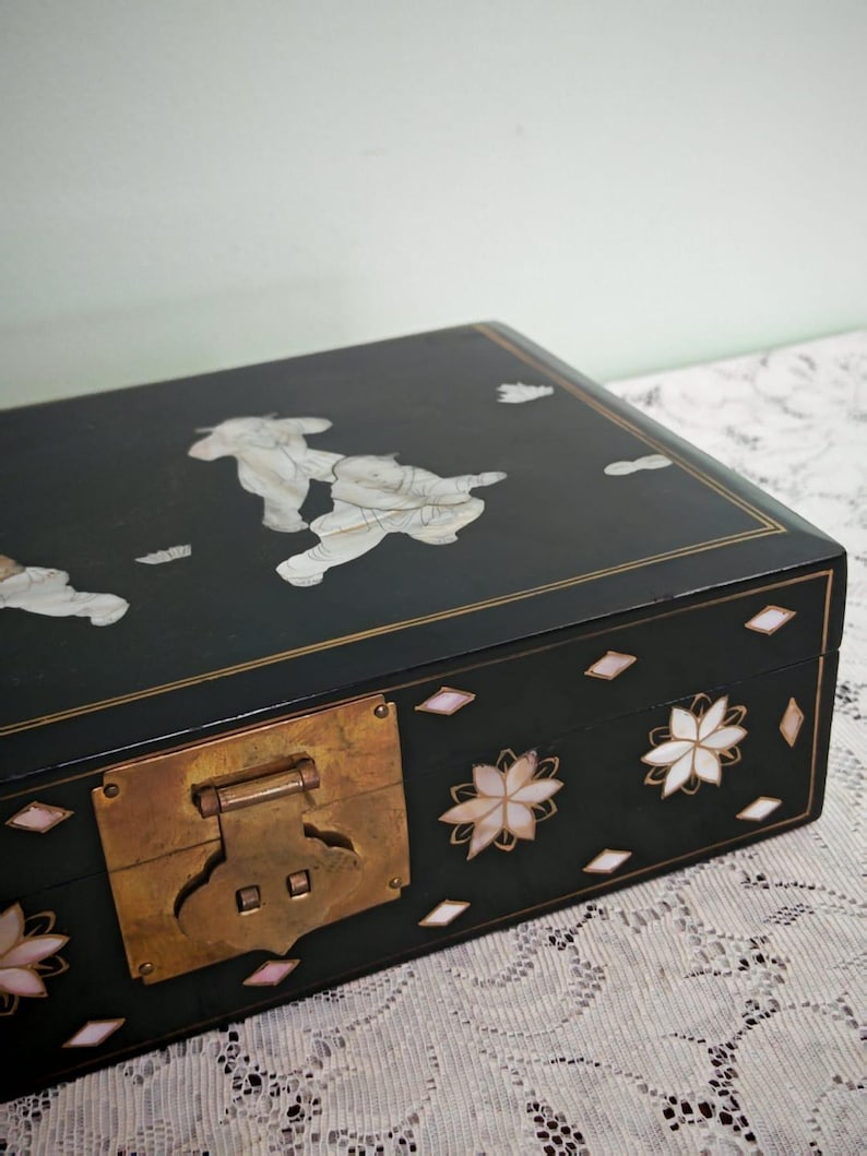 May include: A rectangular black lacquered wooden box featuring inlaid mother-of-pearl figures on the lid and floral motifs on the front. The box includes a prominent brass latch mechanism and thin gold-colored line detailing along the edges.