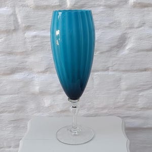 Può includere: Un alto vaso di vetro color teal con un design scanalato. Il vaso ha uno stelo trasparente e attorcigliato e una base rotonda. Lo sfondo è un muro di mattoni bianchi.