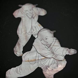 May include: Two flat, iridescent mother-of-pearl inlays shaped like figures in traditional clothing. Each figure features fine, dark engraved lines detailing facial features, hair, and garments. The pieces have a smooth, pearlescent surface texture.