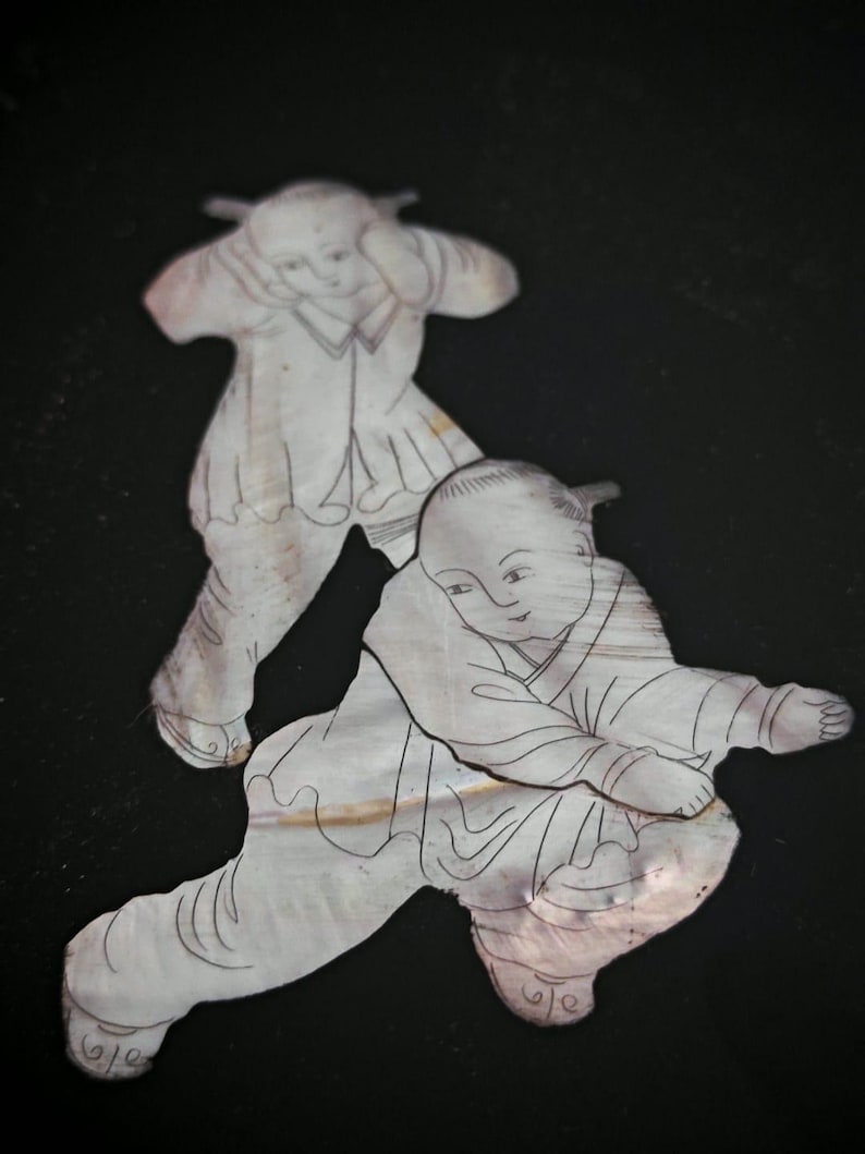 May include: Two flat, iridescent mother-of-pearl inlays shaped like figures in traditional clothing. Each figure features fine, dark engraved lines detailing facial features, hair, and garments. The pieces have a smooth, pearlescent surface texture.