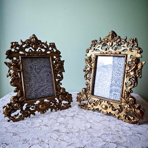 Puede incluir: Dos marcos dorados ornamentados con detalles de querubines y volutas. Cada marco contiene una impresión con un patrón azul y blanco. Los marcos están sobre un mantel de encaje blanco.